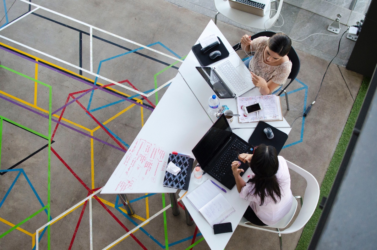Two women sitting in white chairs, talking and using a black and a white laptop on a table.